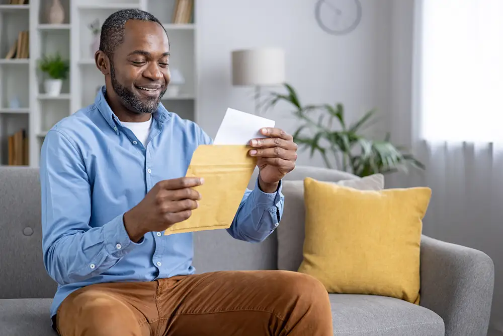 Joyful successful man sitting at home in living room, received mail with notification message, reading contentedly while sitting on sofa.