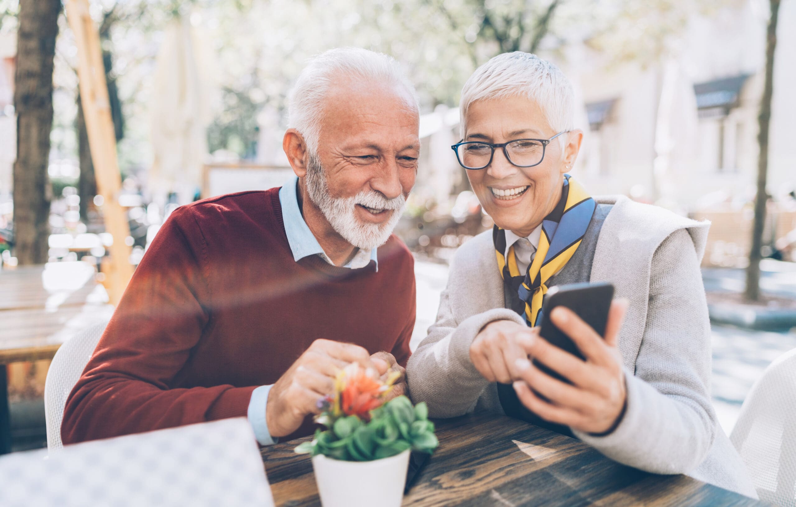 Mature couple with smartphone enjoy Instagram at sidewalk cafe on sunny day