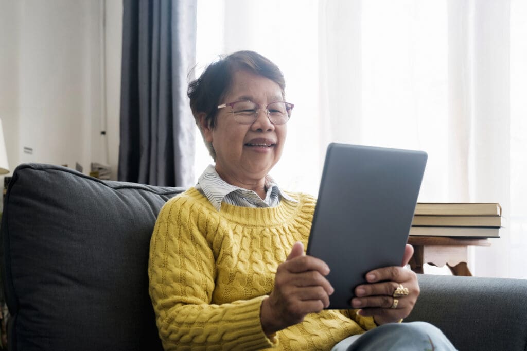 Mature Asian Woman Using Digital Tablet Sitting At Home searching for senior living websites